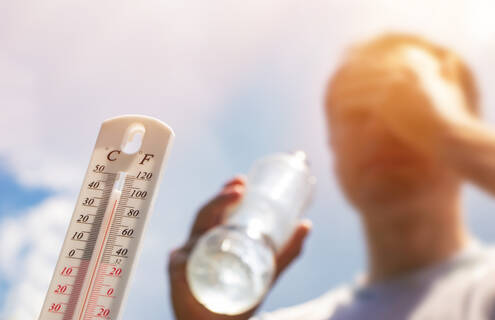 Blurred image of thermometer showing a temperature over 90 degrees Fahrenheit, and a man holding a water bottle and wiping sweat from his face.